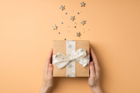 First person top view photo of st valentine's day decorations young female hands holding craft paper giftbox with white ribbon bow glowing stars and star shaped confetti on isolated beige backgroundの写真素材