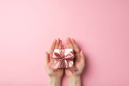 First person top view photo of valentine's day decorations female hands holding small white giftbox with pink ribbon bow on palms on isolated pastel pink background with empty spaceの写真素材