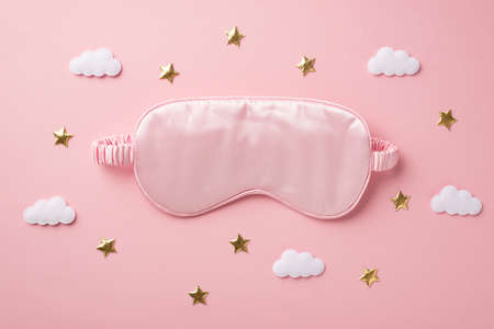 Top view photo of pink silk sleeping mask clouds and golden stars on isolated pastel pink background...