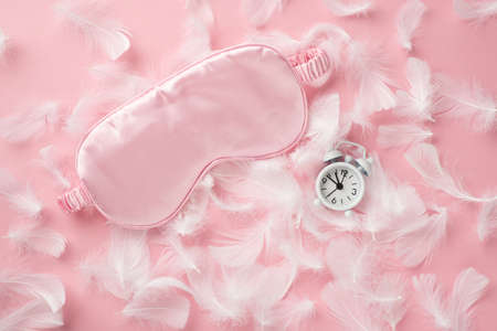 Top view photo of small white alarm clock pink silk sleeping mask and feathers on isolated pastel pink background with empty spaceの写真素材