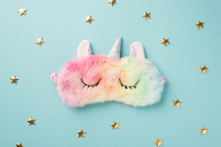 Top view photo of the soft colorful sleep mask in shape of unicorn and cute golden confetti in shape of stars on the isolated pastel blue background blank spaceの写真素材