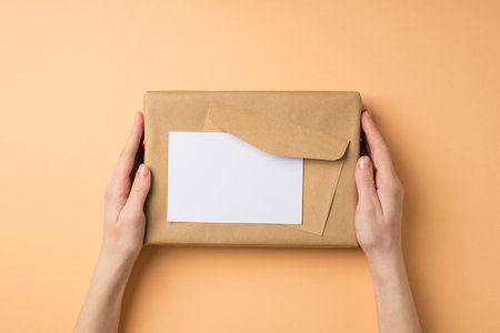 First person top view photo of female hands holding kraft paper giftbox envelope and paper sheet on isolated beige background with blank spaceの写真素材