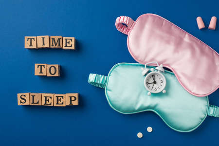 Top view photo of the small white alarm on the two silk masks pink and light blue one with two pills wooden cubes with letters and two pink earplugs on the deep blue backgroundの写真素材