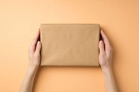 First person top view photo of young woman's hands holding kraft paper giftbox on isolated beige background with empty spaceの写真素材