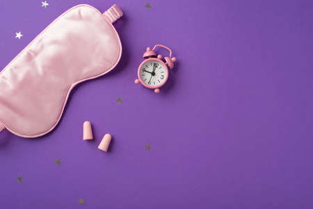 Top view photo of the big silk eye mask and cute pink clock with two earplugs with scattered confetti in shape of stars on the purple isolated background copyspaceの写真素材
