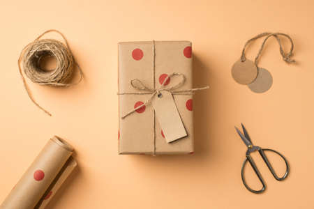 Top view photo of valentine's day decor craft paper giftbox with polka dot pattern tag handicraft tools scissors roll of kraft paper and spool of twine on isolated beige background with copyspaceの写真素材