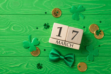 Top view photo of st patrick's day decorations shamrocks wooden cubes calendar 17 march clover shaped confetti green bow-tie and gold coins on isolated textured green wooden desk backgroundの写真素材