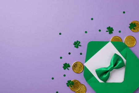 Top view photo of the green envelope with white card inside and silk tie bow on it with golden coins and confetti in shape of shamrocks and dots on isolated empty purple backgroundの写真素材