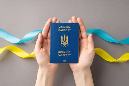 Stop the war in Ukraine concept. Top view photo of female hands holding Ukraine passport on palms over blue and yellow ribbons on gray backgroundの写真素材