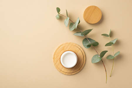 natural cosmetics concept. Top view photo of small cream jar on wooden stand and eucalyptus on isolated beige backgroundの写真素材