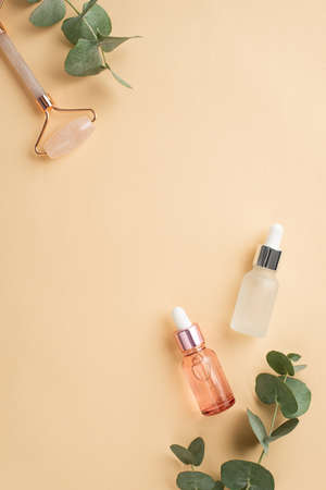 Skin care concept. Top view vertical photo of rose quartz roller two transparent glass dropper bottles and eucalyptus on isolated pastel beige background with empty spaceの写真素材
