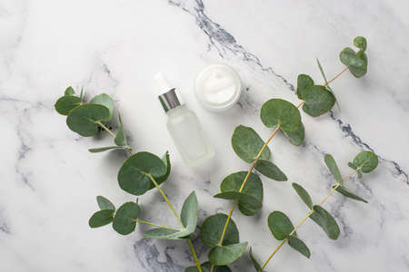 natural cosmetics concept. Top view photo of transparent dropper bottle open cream jar and eucalyptus branches on white marble backgroundの写真素材
