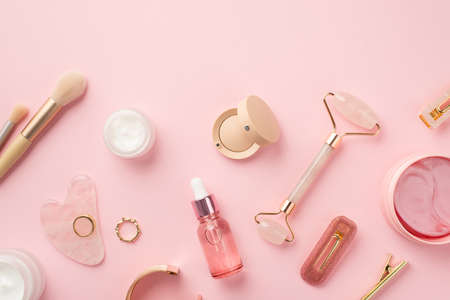 Top view photo of makeup brushes rose quartz roller gua sha pink eye patches glass transparent bottle cream jars eyeshadow barrettes lip gloss gold rings and wristlet on isolated pink backgroundの写真素材