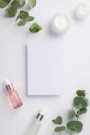 Skin care concept. Top view vertical photo of paper card pink and white glass dropper bottles cream jar and eucalyptus on isolated white background with empty space