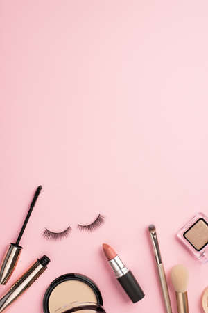 Make up concept. Top view vertical photo of false eyelashes mascara lipstick makeup brushes eyeshadow and compact powder on pastel pink background with copyspaceの写真素材