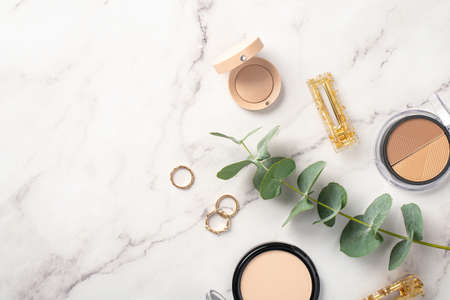 makeup concept. Top view photo of contouring palette eyeshadow compact powder gold rings barrettes and eucalyptus sprig on white marble background with empty spaceの写真素材
