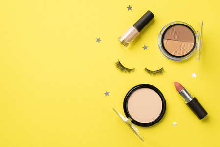 makeup concept. Top view photo of compact powder contouring palette nail polish lipstick false eyelashes and star shaped confetti on isolated yellow backgroundの写真素材