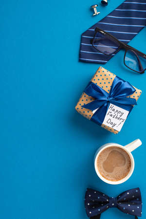 father's day concept. Top view vertical photo of polka dot giftbox with ribbon bow and postcard blue necktie bow-tie glasses cup of coffee and cufflinks on isolated blue backgroundの写真素材