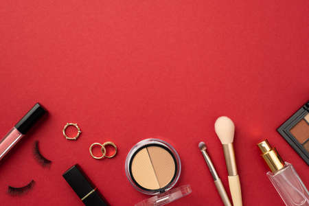 Make-up concept. Top view photo of lip gloss false eyelashes lipstick gold rings contouring eyeshadow palettes makeup brushes and perfume bottle on isolated red background with copyspaceの写真素材