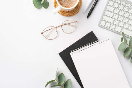 business concept. Top view photo of workplace keyboard black and white stationery note pads pens cup of coffee on rattan placemat glasses and eucalyptus on isolated white background with blank spaceの写真素材