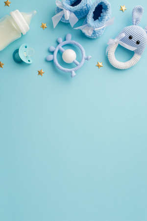 baby boy concept. Top view vertical photo of booties pacifier teether milk bottle knitted bunny rattle toy and gold stars on isolated pastel blue background with copyspaceの写真素材