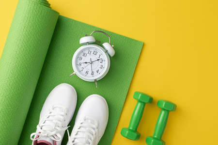 fitness concept. Top view photo of dumbbells white sneakers and alarm clock over green sports mat on isolated yellow backgroundの写真素材