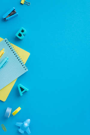 Back to school concept. Top view vertical photo of plastic alphabet letters notebooks pens plane shaped sharpener binder clips adhesive tape and stapler on isolated blue background with empty spaceの写真素材