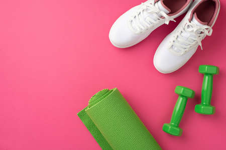 Sports accessories concept. Top view photo of white sports footwear green yoga mat and dumbbells on isolated pink backgroundの写真素材