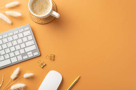 business concept. Top view photo of workspace white keyboard computer mouse gold pen binder clips cup of hot drinking on rattan serving mat and white lagurus flowers on isolated orange backgroundの写真素材