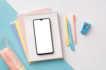 school supplies concept. Top view photo of smartphone over notepads pens pencil-case and mini stapler on bicolor blue and white background with empty spaceの写真素材