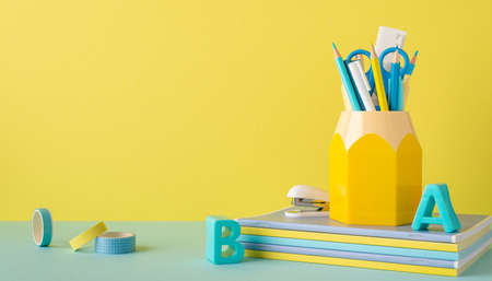 Back to school concept. Photo of school supplies on blue desktop stand for stationery plastic alphabet letters stack of copybooks adhesive tape and stapler on yellow wall background with copyspaceの写真素材