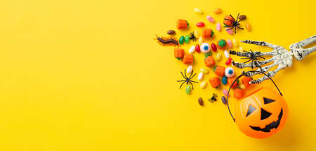 Halloween decoration concept. Top view photo of skeleton hand holding pumpkin basket for trick-or-treat with candies eyes insects centipede and spiders on isolated yellow backgroundの写真素材