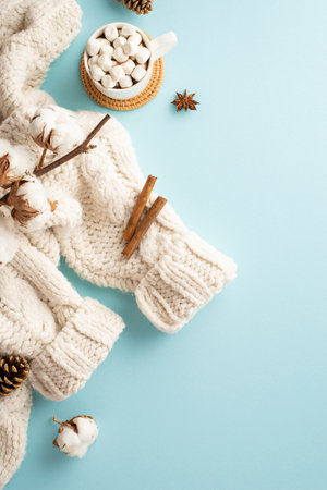 winter concept. Top view vertical photo of white cozy sweater cup of hot drinking with marshmallow on rattan serving mat cotton branch pine cones and cinnamon sticks on isolated pastel blue backgroundの写真素材