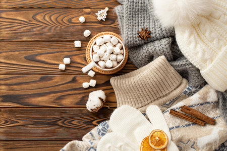 winter concept. Top view photo of cup of cocoa with marshmallow on rattan serving mat jumper bobble hat scarf mittens cotton flower cinnamon sticks dried orange slices anise on wooden desk backgroundの写真素材