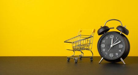 Black friday sales concept. Photo of alarm clock and empty shopping cart on tabletop yellow wall background with copyspaceの写真素材