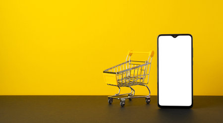 Cyber monday concept. Photo of smartphone and empty shopping cart on tabletop yellow wall background with blank spaceの写真素材