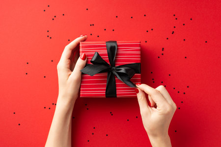 Black friday concept. First person top view photo of female hands unpacking red giftbox with black ribbon bow and confetti on isolated red backgroundの写真素材