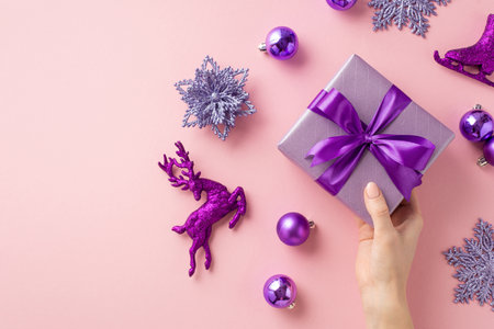 New year concept. First person top view photo of female hand holding lilac present box over violet baubles snowflake flower ice skates deer ornaments on isolated pastel pink background with copyspaceの写真素材