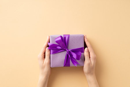 New year concept. First person top view photo of female hands giving purple giftbox with violet ribbon bow on isolated pastel beige background with blank spaceの写真素材