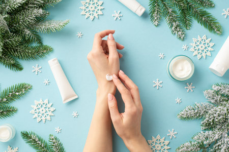 Winter season skin care concept. First person top view photo of woman using hand cream cosmetic jars tubes snowflakes and spruce branches in hoarfrost on isolated pastel blue background with copyspaceの写真素材