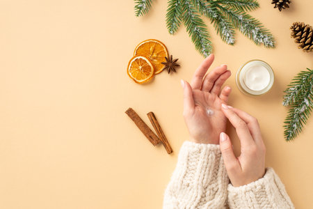 First person top view photo of woman in jumper using hand cream small jar spruce branches dried orange slices pine cone cinnamon sticks and anise on isolated pastel beige background with empty spaceの写真素材