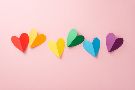 A flat lay top view of LGBT pride items: a rainbow colored paper hearts, placed on a pastel pink...
