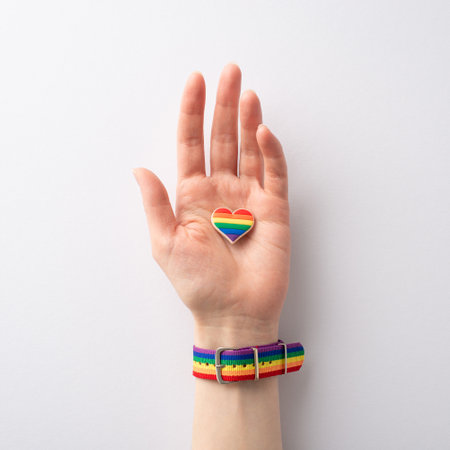 A first person top square view of symbolic rainbow-colored bracelet adorns a female hand holding a heart-shaped pin badge on a white background, in honor of LGBTQ History Monthの写真素材