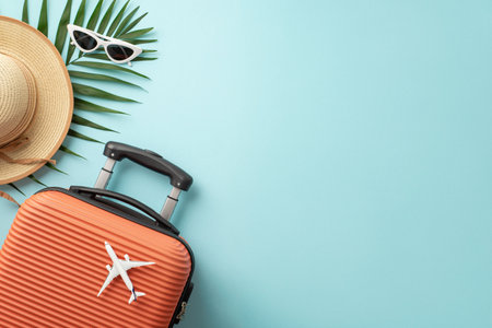 Summer getaway concept. Top view of an orange suitcase, small airplane figurine, beach essentials, sunglasses, sunhat, palm leaves on pastel blue background, providing space for text or promo contentの写真素材