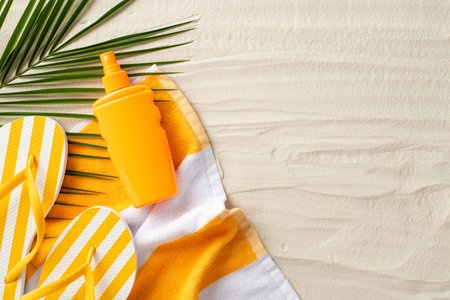 Sun protection on vacation concept. Top view photo of beach towel, yellow flip-flops, sunscreen spray and palm leaf on the sand on isolated background with copyspaceの写真素材