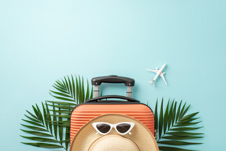 Get ready for summer adventures! Top view of orange suitcase, small airplane model, beach essentials, eyewear, sunhat, and palm leaves on pastel blue background, featuring empty space for text or adsの写真素材