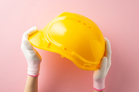 Tribute to hardworking women on Labor Day. First person top view of woman's gloved hands with safety helmet on pink backdrop. Advertise hereの写真素材