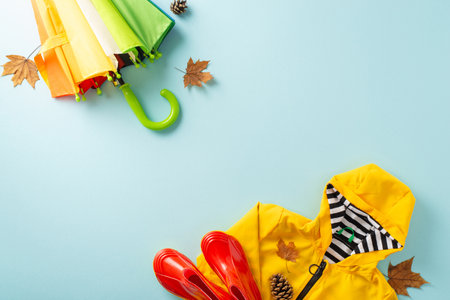 Seasonal serenity. Top view scene featuring child's yellow raincoat, red gumshoes, vibrant umbrella with rainbow pattern, maple leaves, cones on pastel blue backdrop, providing space for text or adの写真素材