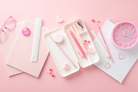 A desk full of inspiration. Top view of school supplies and stationery on a pastel pink backdrop. Pencil case, pens, stapler, ruler, glitter stone, album, heart shaped clips and more, ideal for adsの写真素材