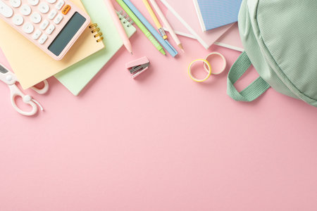 Modern girl's educational gear display. Top view flat lay arrangement featuring rucksack, stapler, tape, scissors, copybooks, calculator on pastel pink backdrop, with empty space for text or promotionの写真素材